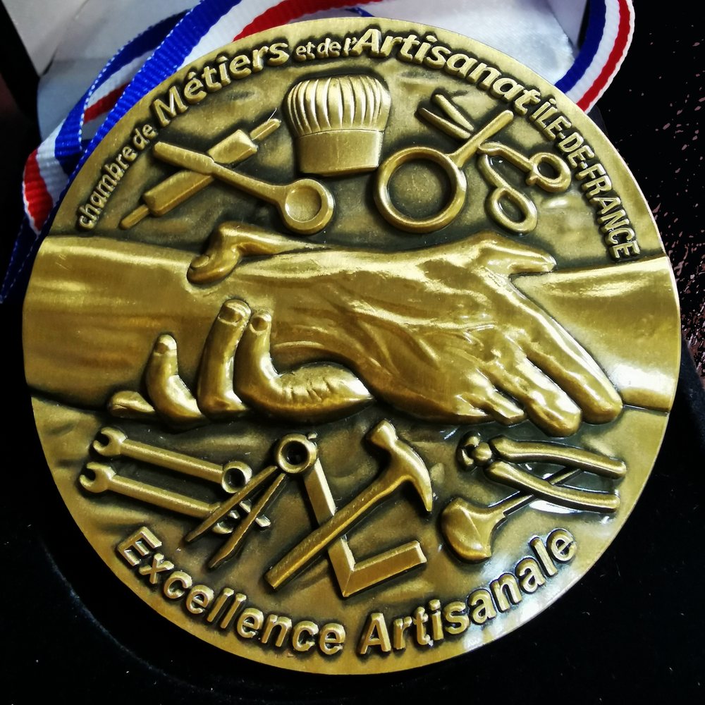 Médaille de l'Excellence artisanale : deux mains qui se serrent, entourées des outils de l'artisan